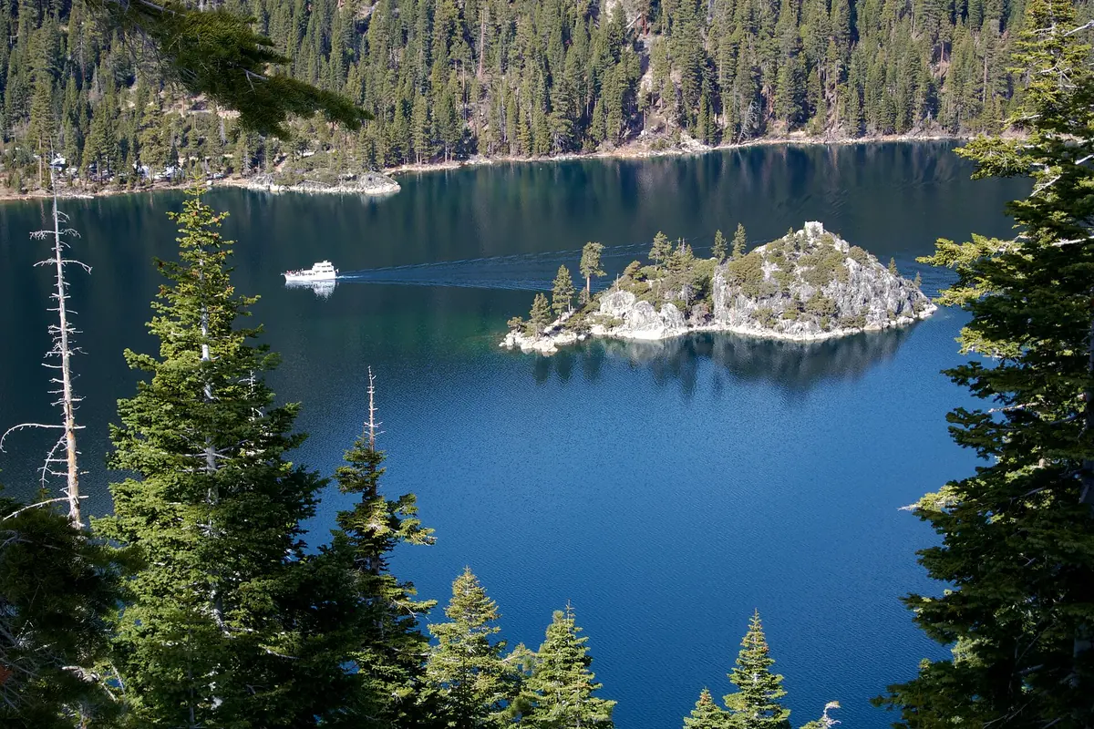 Fannette Island, a small rocky island with pines, in the deep blue water of Emerald Bay, with a white boat passing and forested slopes behind